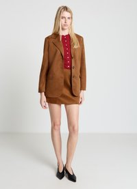 Blazer en daim marron avec revers à encoches, trois boutons et deux poches avant, associé à une chemise rouge boutonnée et une mini-jupe marron. Talons noirs.