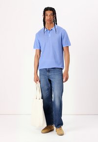 Lyseblå kortermet polo med et teksturert mønster, parret med blå jeans. Modellen holder en kremfarget totebag og har på seg beige sneakers.