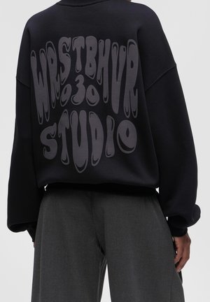 Personne portant un sweat-shirt noir surdimensionné avec un grand texte déformé « WRSTBHVR 30 STUDIO » dans le dos, assorti à un pantalon gris foncé.