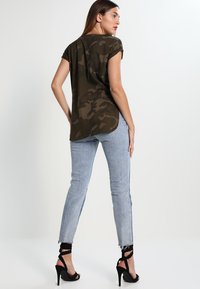 Camo-patterned olive green top with a rounded hem, paired with light blue jeans featuring distressed details and back ankle strap heels.