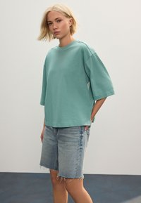 Tricou supradimensionat de culoare teal deschis, din bumbac moale, cu decolteu rotund și mâneci scurte, asortat cu pantaloni scurți din denim cu margini franjurate.