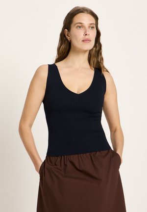 Frau mit langem, welligem Haar, die ein schwarzes, ärmelloses, geripptes Oberteil und einen braunen Rock trägt, steht mit den Händen in den Taschen vor einem einfarbigen Hintergrund.