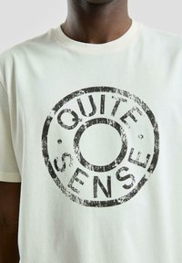Zwart versleten tekst "QUIET SENSE" in een cirkelvormig stempelontwerp op een witte t-shirt met ronde hals dat door een persoon wordt gedragen.