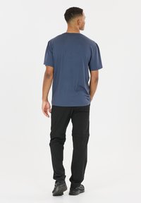 Mand set bagfra iført en blå kortærmet skjorte, sorte konvertible bukser og sorte vandresko mod en hvid baggrund.