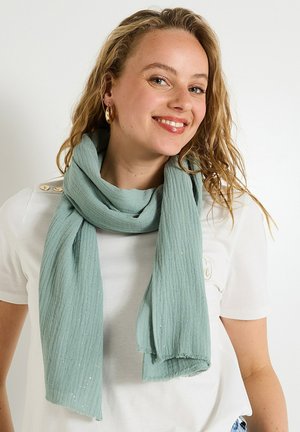 Écharpe bleu sarcelle légère avec un motif texturé en côtes et des éléments de brillance subtils. Les bords sont effilochés. Portée lâchement autour du cou.