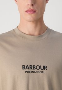 Beez värvi puuvillane T-särk, millel on ümmargune kaelus ja must trükitud tekst "BARBOUR INTERNATIONAL" rinnal.