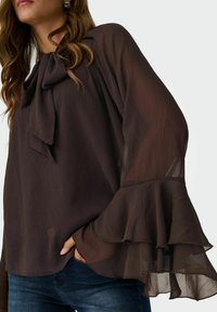 Femme portant un chemisier marron foncé transparent avec des manches cloche superposées et un col à nouer, assorti à un jean bleu.