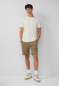 Jasno beżowy t-shirt z okrągłym dekoltem, zestawiony z khaki szortami. Model nosi białe skarpetki i beżowe sneakersy z granatowymi akcentami.
