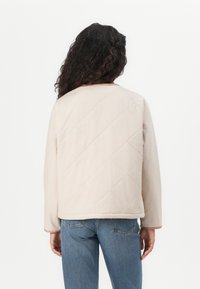 Beige quiltad jacka med ett rutmönster, rund halsringning och långa ärmar, i kombination med ljusblå jeans, synlig bakifrån.