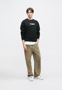 Μαύρη μπλούζα με λογότυπο, στρωμένη πάνω από λευκό μπλουζάκι. Το μοντέλο φοράει μπεζ παντελόνι φαρδιάς γραμμής και λευκά αθλητικά παπούτσια. Απλό φόντο.