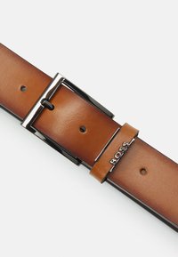 Curea din piele maro cu o cataramă argintie lustruită. Include un bucla asortată cu numele brandului embosat. Dispune de mai multe găuri pentru ajustare.