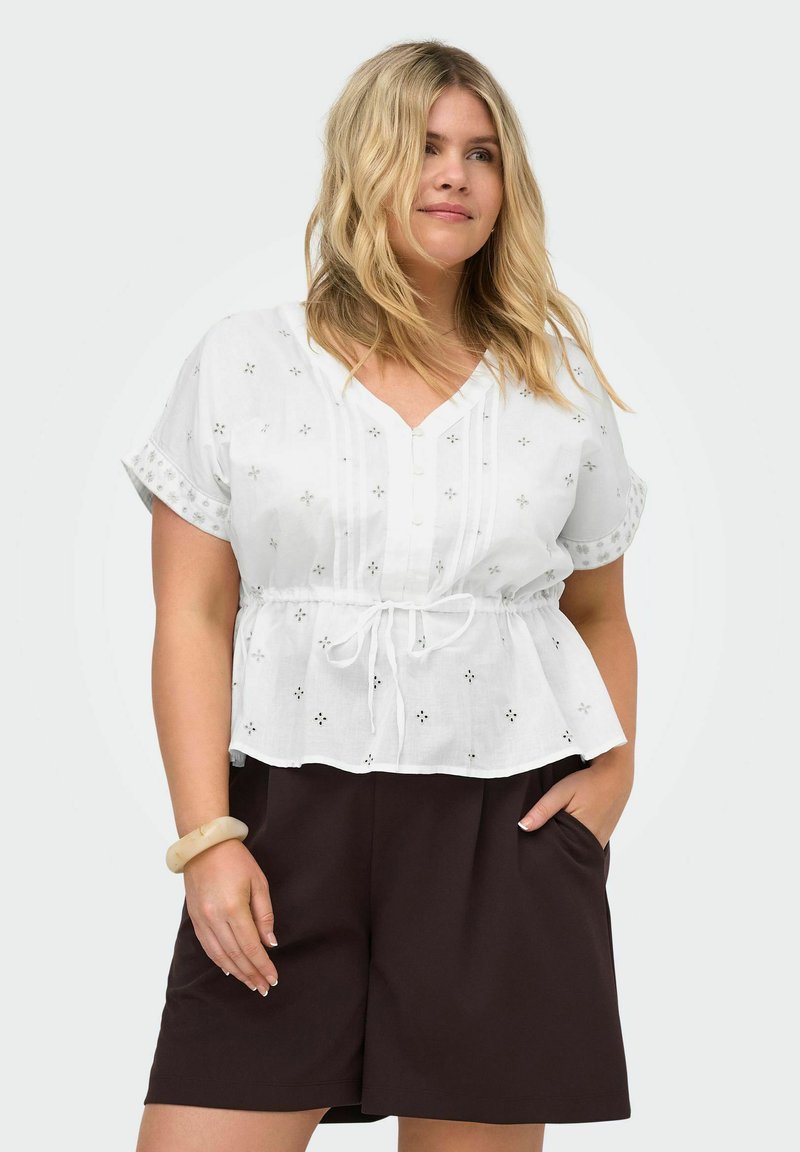 Femme blonde portant un chemisier blanc à manches courtes avec des détails ajourés et un short noir ample, main dans la poche, et un bracelet beige.