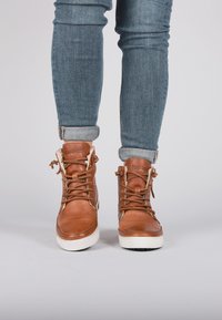 Botas de tobillo de cuero marrón con superficie texturada, suela de goma blanca, punta redonda y ojetes metálicos. Presenta cordones y forro de felpa.