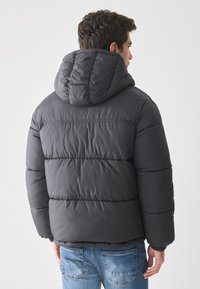 Hombre vestido con una chaqueta acolchada gris oscuro con capucha, de pie de espaldas, acompañado de jeans de mezclilla azules, contra un fondo blanco liso.
