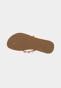 Επίπεδη σόλα σανδάλι σε καφέ λάστιχο με υφασμένο κράτημα, λουριά σε ροζ χρυσό και μεταλλικές διακοσμήσεις, απεικονιζόμενο από κάτω.