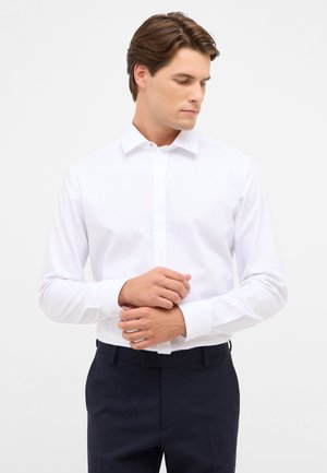 Jonge man die de manchet van een wit overhemd aanpast, gedragen met een donkere broek, terwijl hij naar beneden en opzij kijkt tegen een effen lichte achtergrond.