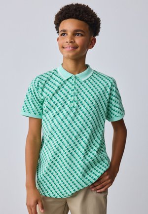 Dreng med krøllet hår iført en mintgrøn mønstret polo og beige bukser, smilende og kigger let til siden.