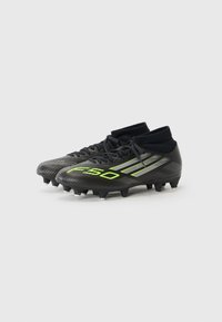 Zwarte voetbalschoenen met groene accenten en een hoog enkelontwerp, geplaatst op een lichtgrijze achtergrond.