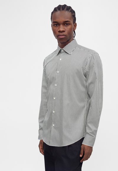 Homme aux cheveux tressés portant une chemise blanche à boutons avec de fines rayures verticales noires et un pantalon noir, debout devant un fond uni.