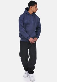 Mørkeblå hættetrøje med et stort logo i en mørkere nuance, parret med sorte bukser og hvide sneakers. Hættetrøjen har en frontlomme og snørelukning.