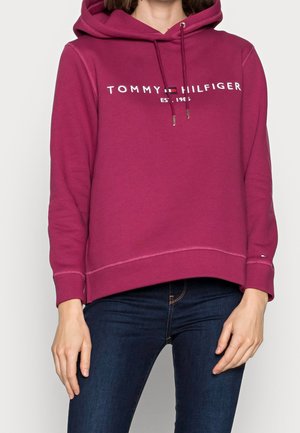 Vrouw draagt een donkeroze Tommy Hilfiger capuchontrui met logo en donkerblauwe jeans, staat met ontspannen armen langs haar zij.
