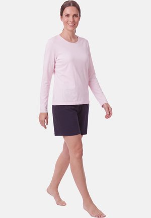 Glimlachende vrouw loopt blootsvoets en draagt een lichtroze shirt met lange mouwen en donkere knielange shorts op een effen witte achtergrond.
