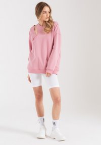 Różowa, oversize'owa bluza z detalem wycięcia, w parze z białymi, prążkowanymi szortami rowerowymi i białymi sneakerami. Skarpetki mają napis "Do zobaczenia później."