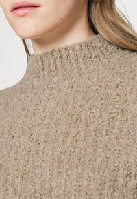 Pull beige en tissu bouclé texturé. Caractéristiques d'un col montant côtelé et une apparence douce et confortable. Vue rapprochée du matériau.