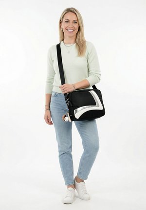 Kvinde smilende, stående, iført lys sweater og jeans, bærende på sort og sølvfarvet skuldertaske med nøgleringsaccessories.