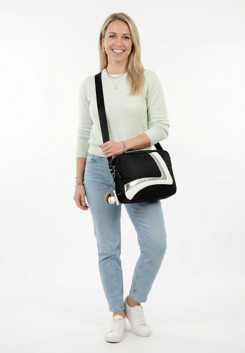 Femme souriante, debout, portant un pull léger et un jean, avec un sac à épaule noir et argenté orné de porte-clés.