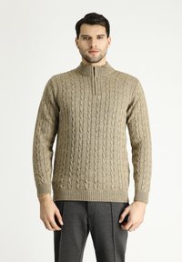 Beiger Strickpullover mit Kabelmuster, hohen Kragen und gerippten Bündchen und Saum. Getragen mit grauen Hose, die eine lockere Passform zeigt.