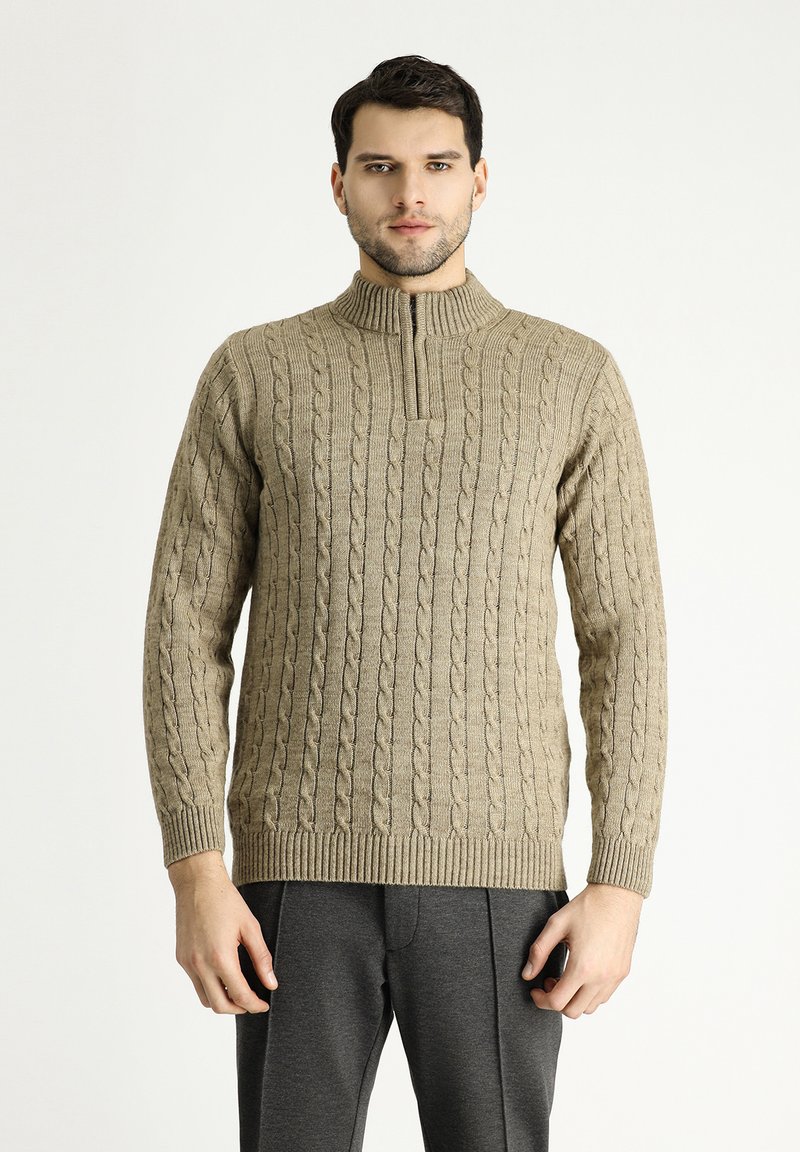 Beiger Strickpullover mit Kabelmuster, hohen Kragen und gerippten Bündchen und Saum. Getragen mit grauen Hose, die eine lockere Passform zeigt.