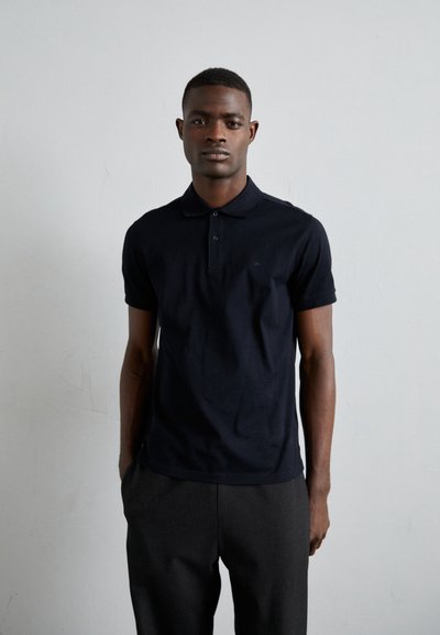 Polo shirt marine à manches courtes avec patte de boutonnage à deux boutons, associé à un pantalon gris foncé. Texture de tissu lisse, branding minimal.