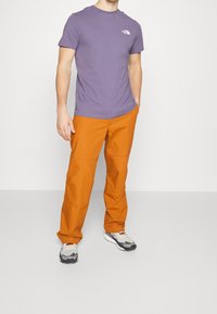 Lila bomullströja med rund hals och liten vit logotyp; orange slitstarka byxor med fickor, tillsammans med gråa och svarta träningsskor.