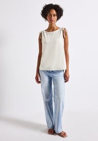 A woman with curly hair stands in a light-colored top and blue jeans, posing against a plain white background.