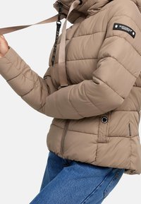 Beige gewatteerd jack met ritssluiting, een verstelbare capuchon en een logopatch op de mouw; heeft horizontale stiksels en handzakken.