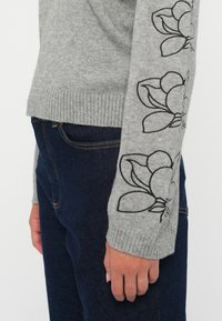 Gråstrikket genser med svart blomsterbroderi på ermene, ribbestrikkede mansjetter og en avslappet passform, kombinert med mørkeblå denim.
