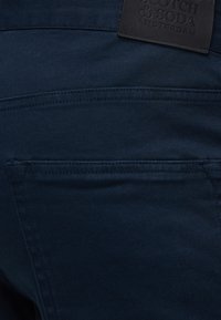 Πίσω τσέπη σκούρου μπλε παντελονιού με ορατές ραφές και μαύρη δερμάτινη ετικέτα με ανάγλυφο το κείμενο "Scotch & Soda Amsterdam".