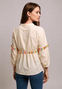 Blusa color crema con mangas tres cuartos, que presenta un borde de borlas coloridas a lo largo de la cintura, confeccionada en una tela ligera.