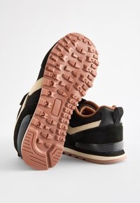 Baskets noires avec une tige texturée, des accents marron clair, une semelle extérieure en caoutchouc avec des crampons profonds, et un talon coussiné orné d'une bande grise.