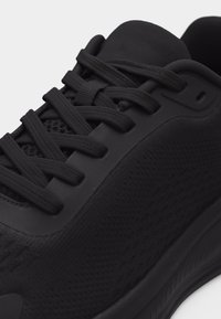 Μαύρο αθλητικό παπούτσι με ύφασμα από διχτυωτό υλικό, χοντρή σόλα και μαύρα κορδόνια σφιχτά δεμένα πάνω σε επενδεδυμένη γλώσσα.