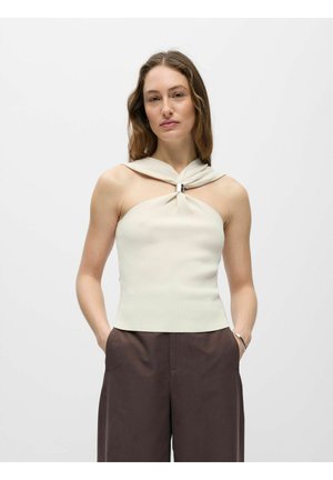 Femme portant un haut beige sans manches avec un détail anneau argenté et un pantalon marron foncé taille haute, debout les mains dans les poches.