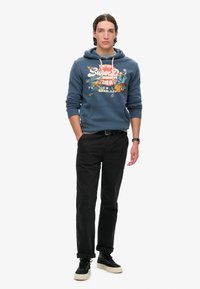 Sudadera azul marino con diseño gráfico, capucha con cordón y bolsillo frontal. Combinada con pantalones negros y zapatillas negras con acentos crema.