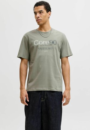 T-shirt en coton vert olive avec l'inscription "Core90" imprimée en texte blanc et gris. Manches courtes, coupe classique, avec un col rond.