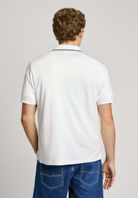 Balta kokvilnas polo krekls ar klasisku apkakli, īsām piedurknēm un smalkām jūras zilām akcentiem apkakles malā, kopā ar ziliem džinsu šortiem.