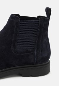 Gros plan d'une botte Chelsea en daim bleu marine avec un panneau latéral élastique texturé et une semelle en caoutchouc noir, avec une languette à l'arrière.