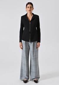 Blazer noir ajusté avec une fermeture éclair à l'avant et un col pointu, associé à un pantalon à carreaux gris à jambes larges et des chaussures noires à enfiler.