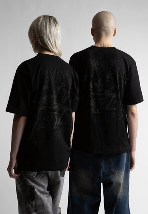 Dwie osoby zwrócone tyłem noszą czarne koszulki z nadrukiem; jedna ma długie blond włosy i podniszczone szare jeansy, druga jest łysa i ma niebieskie jeansy.