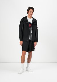 Sort letvægtsjakke med hætte, lynlåslommer og kontrasterende hvid syning, parret med sorte shorts og hvide sneakers.