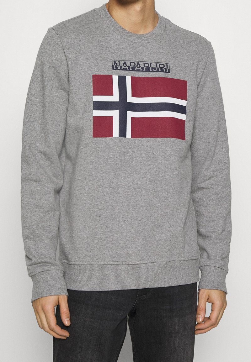 Hombre que lleva un sudadera gris con una bandera con la cruz nórdica en rojo, blanco y azul, y el nombre de la marca "Napapijri" en el pecho.